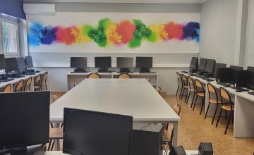 Odnowiona sala lekcyjna. Pod biały ścianami stoją biurka z komputerami. W centralej częsci ustawiony jest biały stół z krzesłami.
